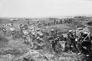 WWI soldiers march in line next to a line of cannons with an earlier tank in the background