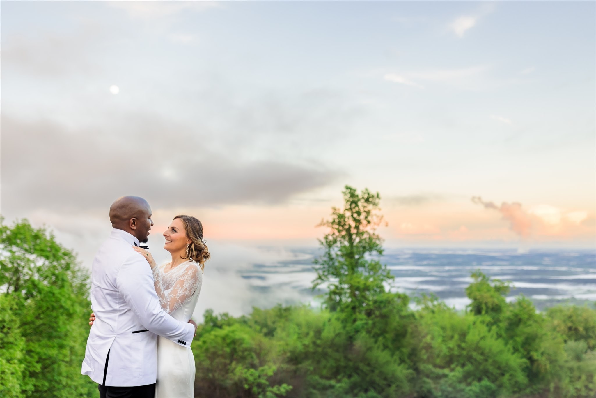A bride and groom pose with a sunset, mountaintop view in the background