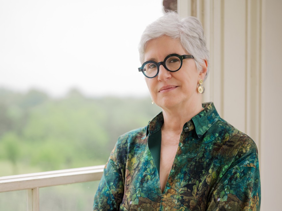 Headshot of Kamensky, in a green floral-patterned shirt and round green-rimmed glasses, posing next to an open window