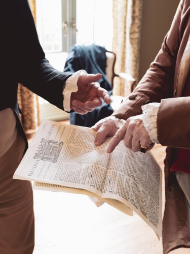 The hands of two men dressed in early 19th century garb gesture at a newspaper