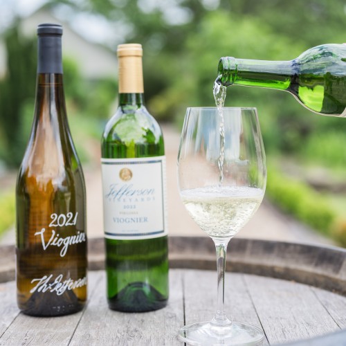 A bottle of Jefferson Vineyard white wine is poured into a glass on the flat end of an upright wine barrel with other signature wines on display next to the glass