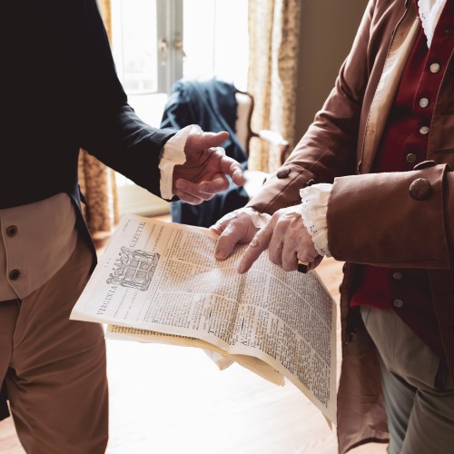 Two historical re-enactors, visible only from the shoulders down, from the late 18th/early 19th century hold a newspaper and gesture energetically