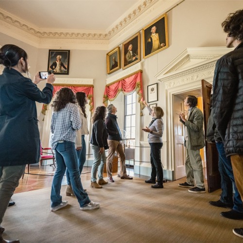 Someone giving a house tour as group of visitors to look up at paintings of Thomas Jefferson, John Adams, and George Washington.