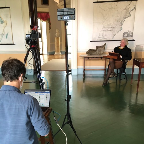 Thomas Jefferson (an interpreter) sits in a chair while a videotechnician works on a computer behind a camera