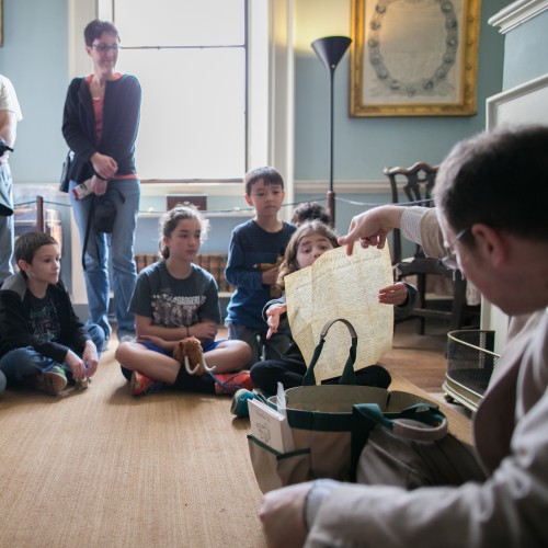 A guide and a group of children sit on the floor while a guide giving a tour holds up a copy of the Declaration of Independence on Monticello's Family Friendly Tour